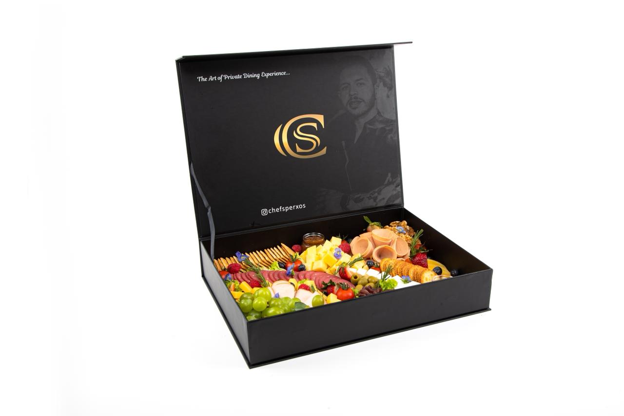 An open black gourmet box filled with neatly arranged fruits, cheese, and snacks